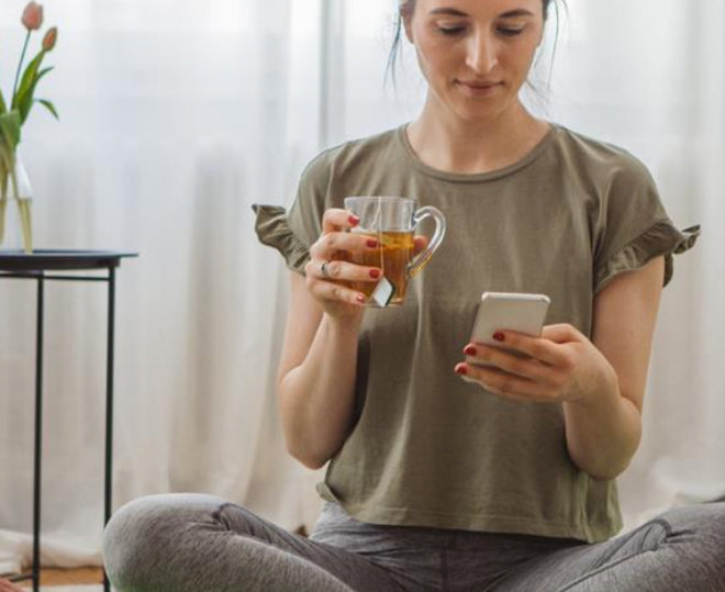 Woman scrolling through her iphone while drinking a cup of avocado leaf tea.
